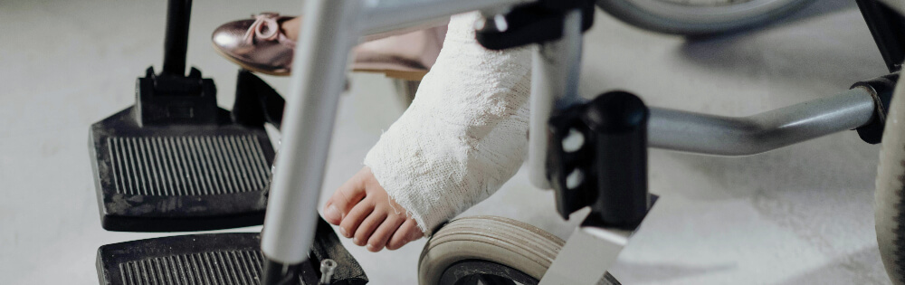 Nahaufnahme eines Fußes in einem Gipsverband, der auf der Fußstütze eines Rollstuhls ruht. Der andere Fuß trägt einen rosa Schuh. Der Rollstuhl hat schwarze Fußstützen und silberne Rahmen.